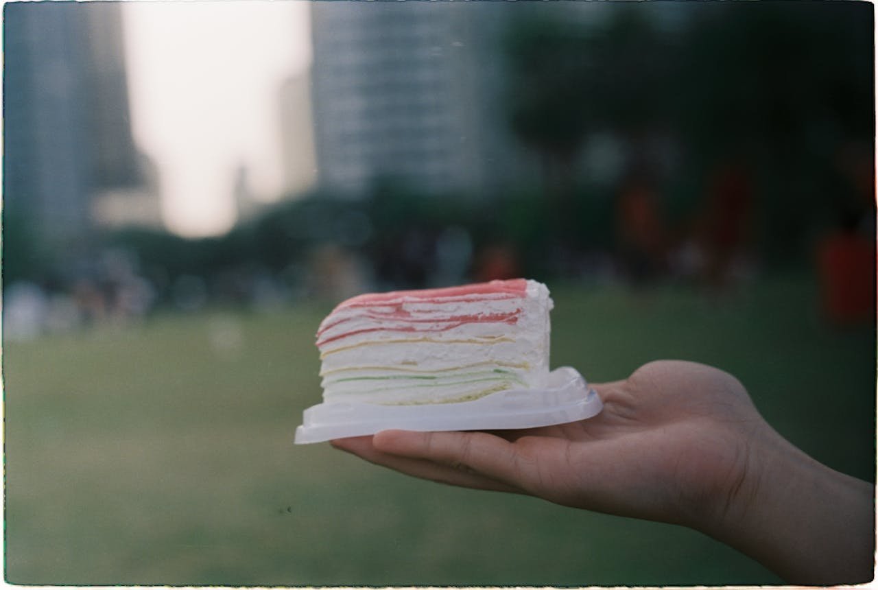 A hand holding a multicolored cake slice outdoors with blurred cityscape.
